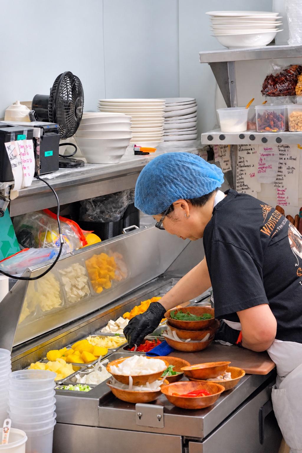 A cook in a blue hairnet and black gloves at the cold prep station, filling wooden bowls with onion, chilli, basil and pineapple from the pans in front of her.